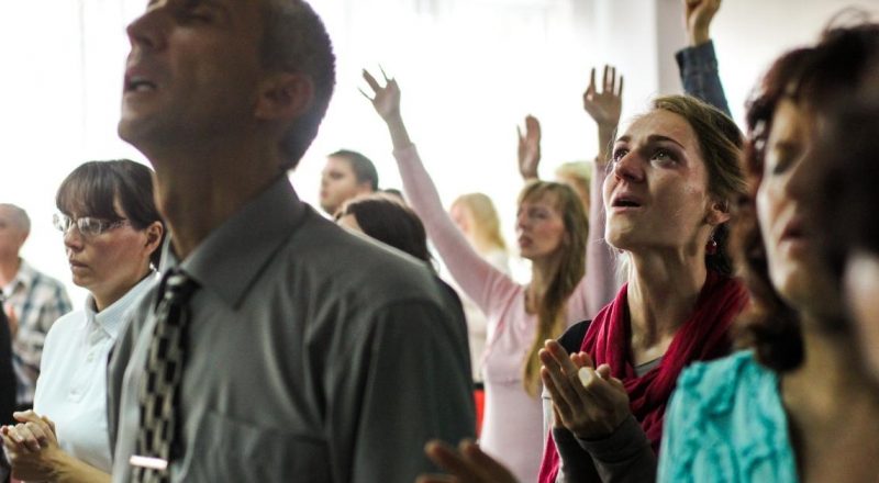A group of people in a church service who are worshipping with their hands up and eyes closed. They appear to be singing.